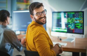 Become a software developer concept. Smiling, bearded software developer looks at camera over shoulder with colleague in background