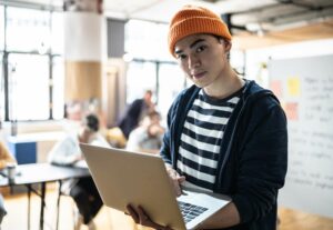 IT staff augmentation concept. Young man with orange beany hat holding an open laptop.